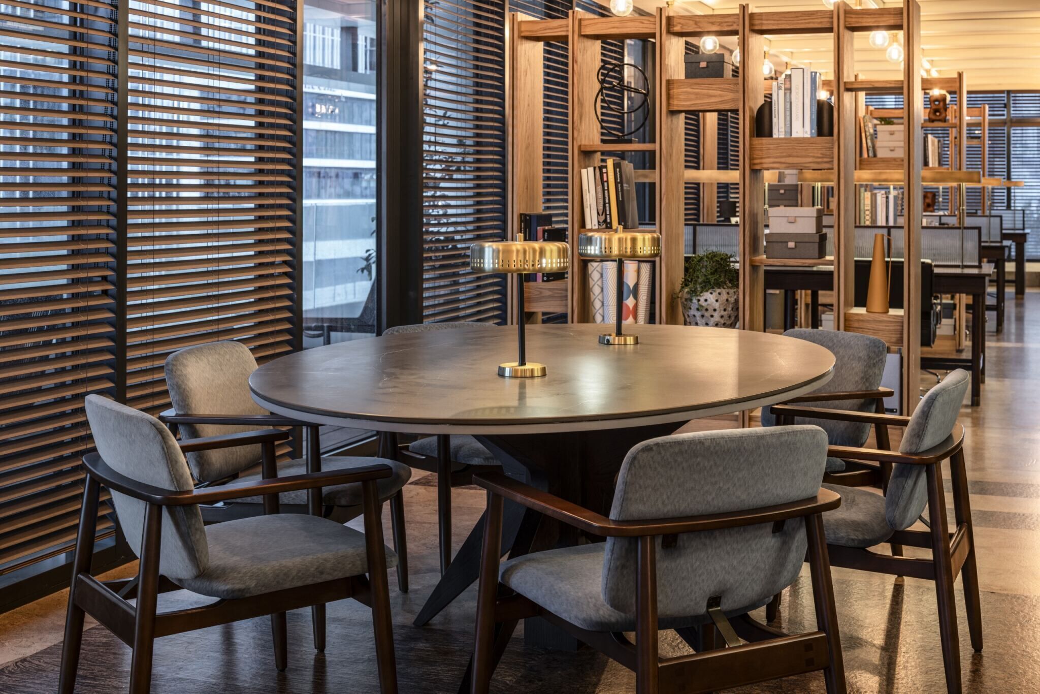 A round table with six chairs, a wooden bookshelf, and a large window with blinds.