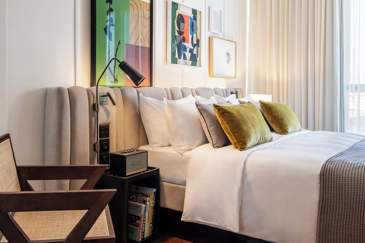 A modern bedroom with a bed, a chair, a bookshelf, and artwork on the wall.
