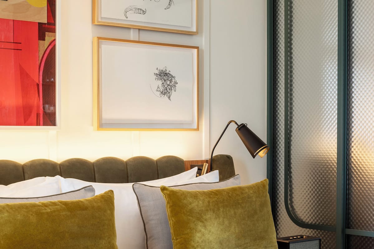 A neatly made bed with a white comforter and two mustard-colored pillows, a bedside lamp, and two framed artworks on the wall.