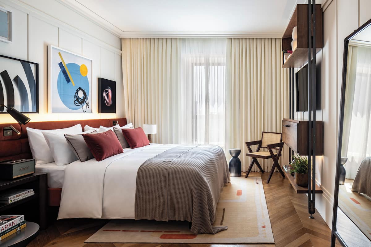 A modern hotel room with a large bed, a desk, a chair, a TV, and artwork on the walls.