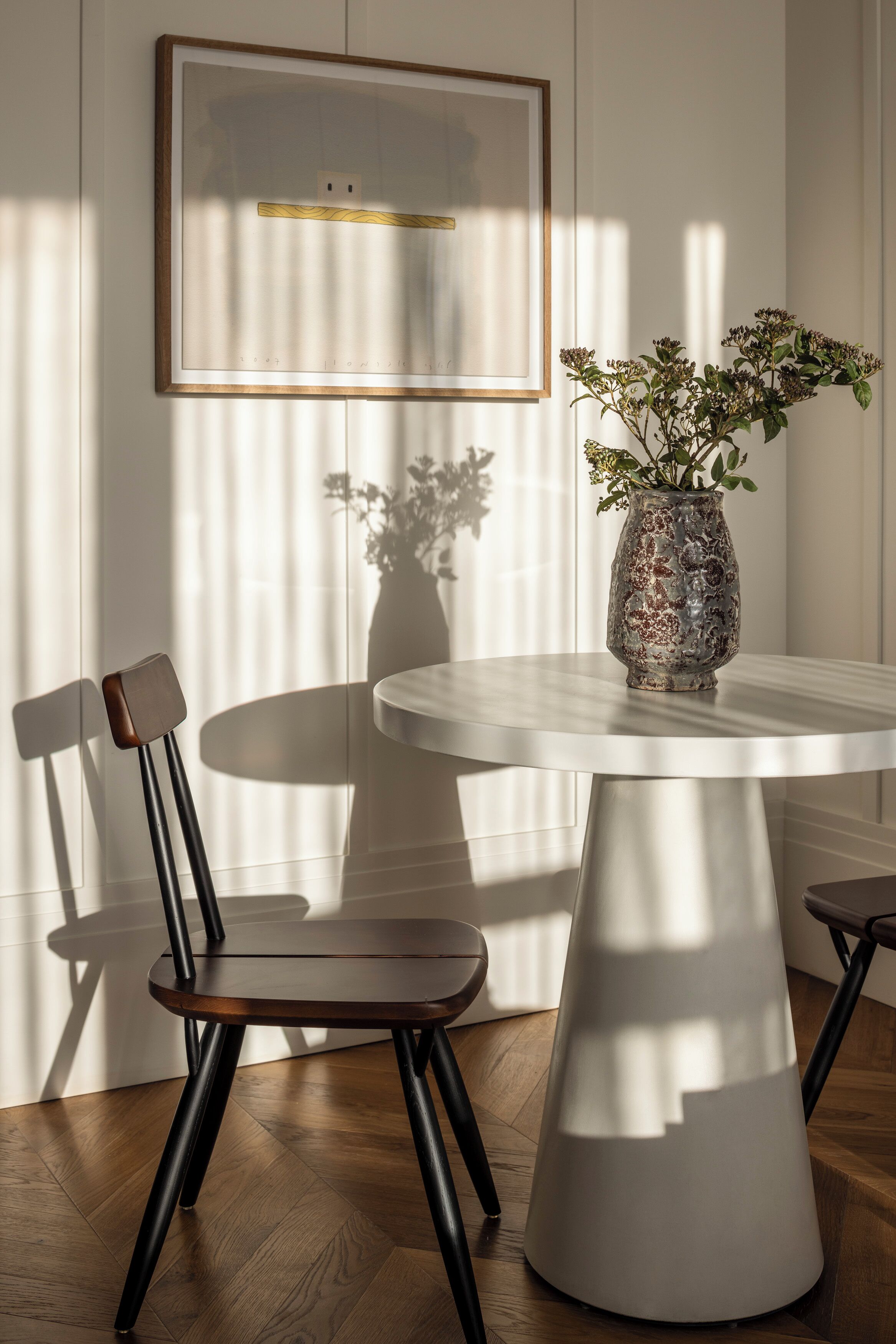 A dining area with a round table, two chairs, a vase with flowers, and a framed picture on the wall.