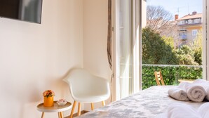 Une chambre avec un lit, une petite table sur laquelle est posé un vase de fleurs, une chaise blanche et une vue sur des immeubles et des arbres, visible par la fenêtre.
