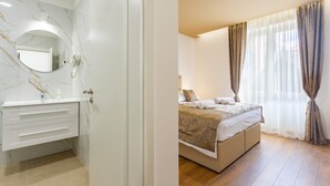 Une chambre d’hôtel dotée d’un meuble-lavabo en marbre dans la salle de bain, d’un lit recouvert d’une couvre-lit à motifs beige et marron, et d’une grande fenêtre agrémentée de rideaux légers.