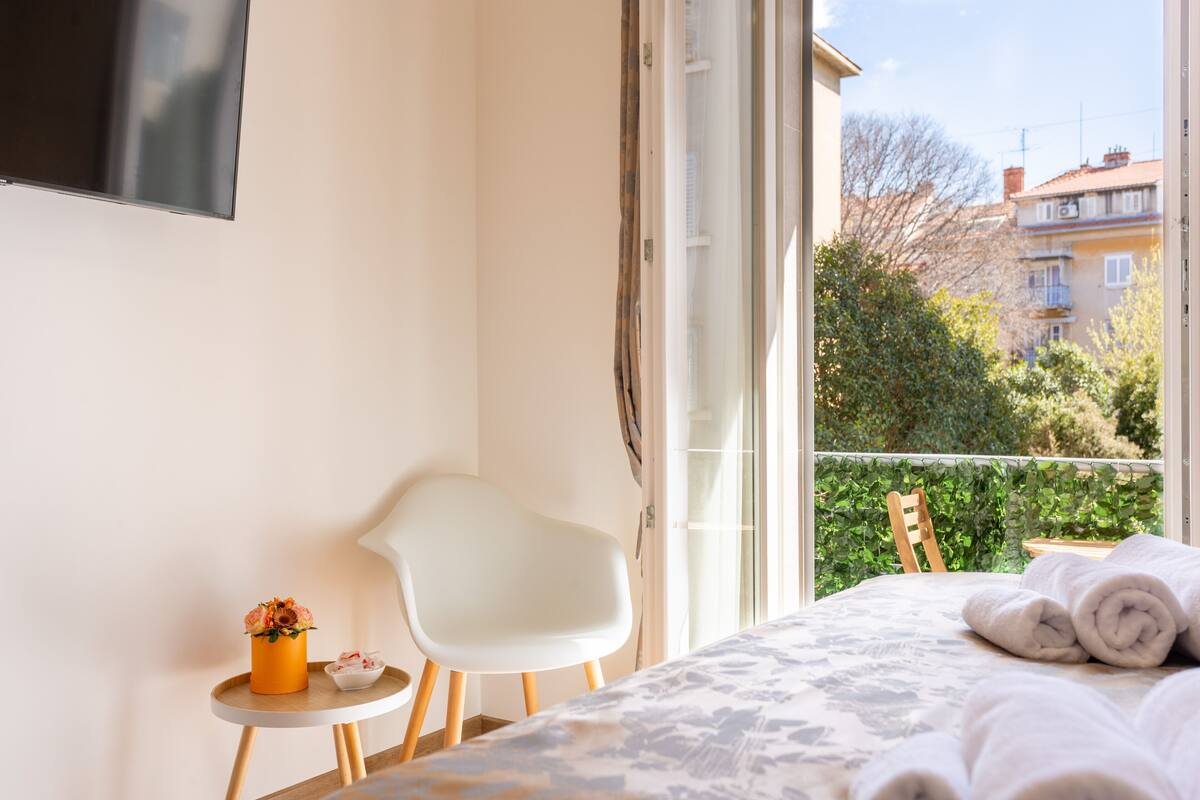 A bedroom with a bed, a chair, a small table with a vase of flowers, and a view of buildings and trees through a window.