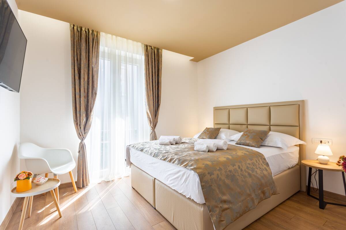 A modern hotel room with a large bed, a bedside table with a lamp, a chair, and a TV mounted on the wall.