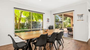 Ein moderner Essbereich mit einem Holztisch und schwarzen Stühlen, großen Fenstern mit Blick auf Grünflächen und einer Glasschiebetür, die zu einer Außenpatio führt.