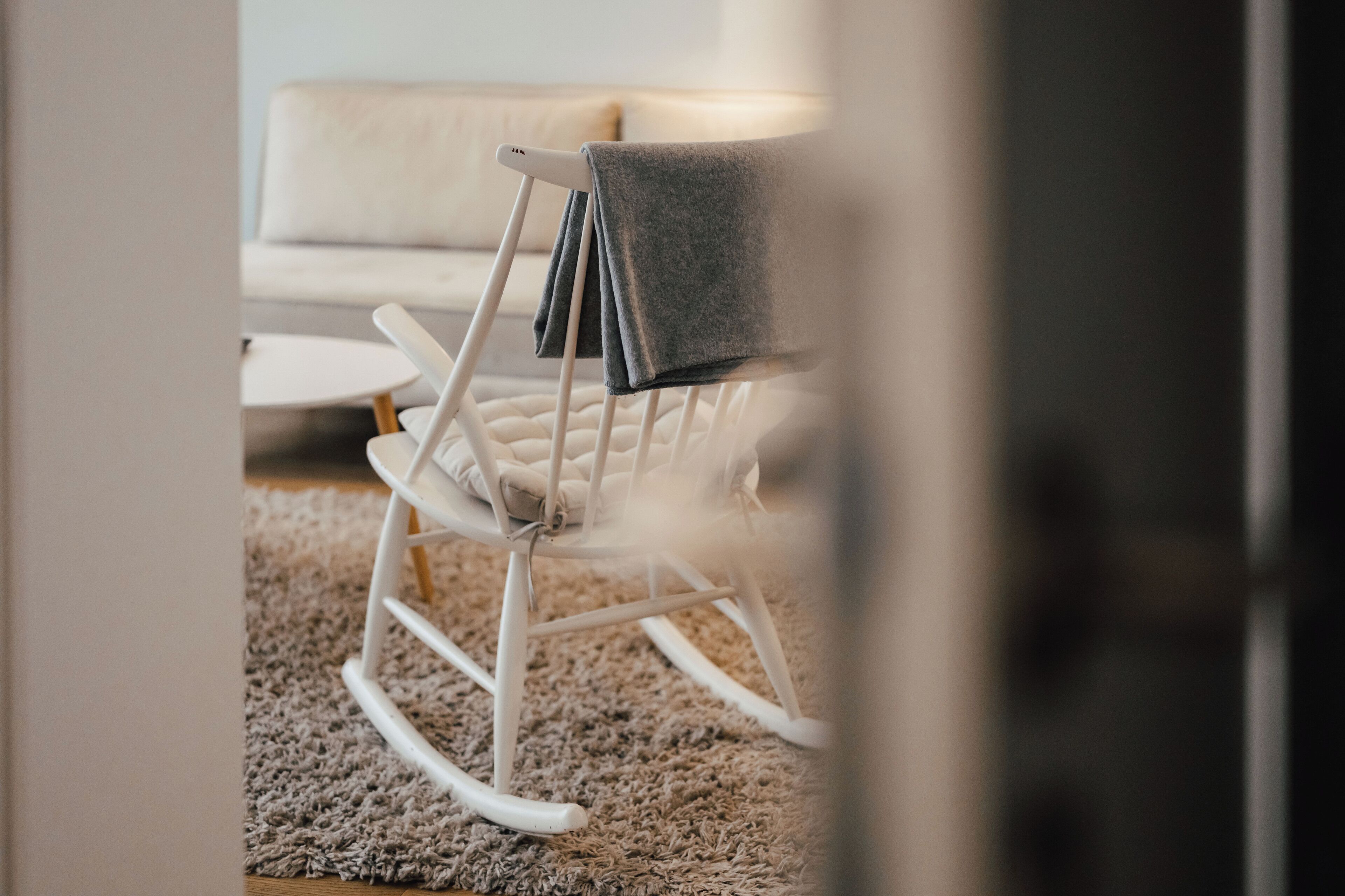 A white rocking chair with a grey blanket, a white sofa, and a textured carpet in a room.