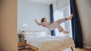 A person jumping on a bed in a room with a wooden floor, a bedside table with a vase of flowers, and a window with curtains.