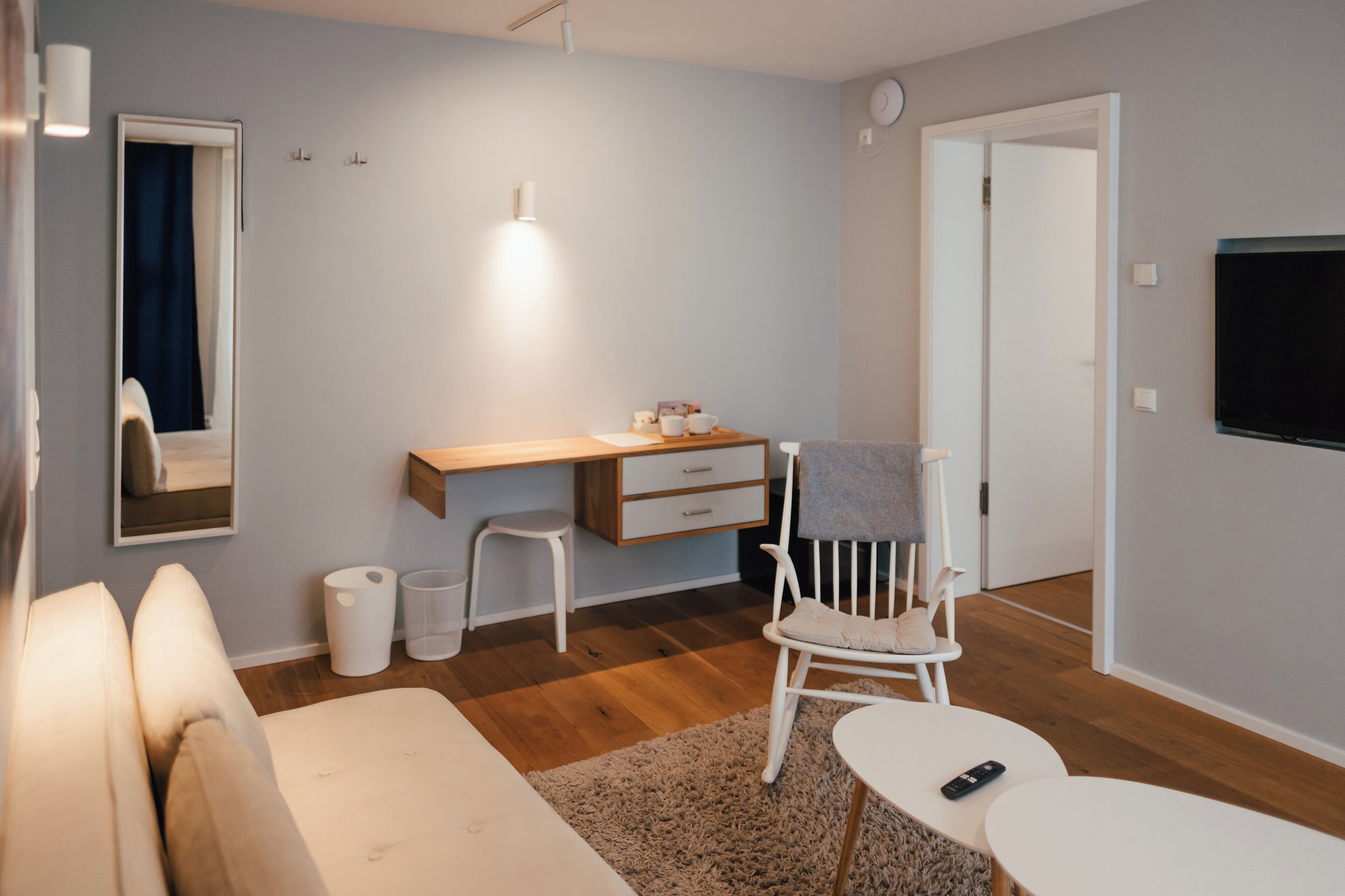 Ein modernes, helles Zimmer mit Holzboden, einer weißen Couch, einem Schreibtisch mit Stuhl, einem kleinen Tisch und einem an der Wand befestigten Fernseher.