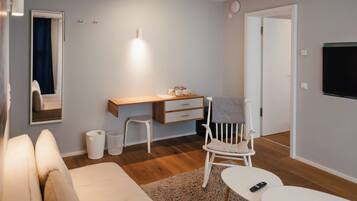 Ein modernes, helles Zimmer mit Holzboden, einer weißen Couch, einem Schreibtisch mit Stuhl, einem kleinen Tisch und einem an der Wand befestigten Fernseher.