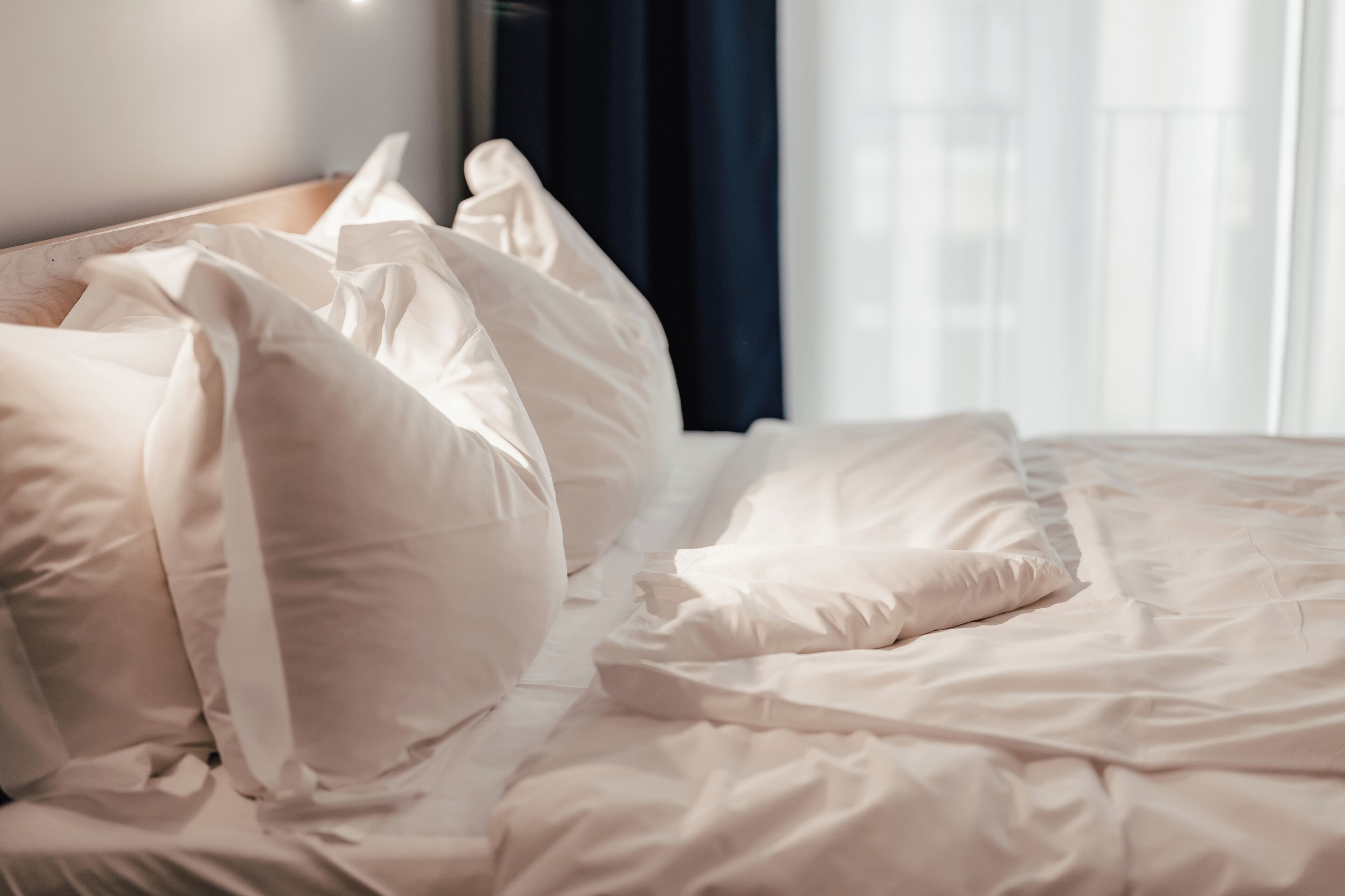 Una cama bien tendida con sábanas y almohadas blancas, cabecera de madera y una ventana con cortinas.
