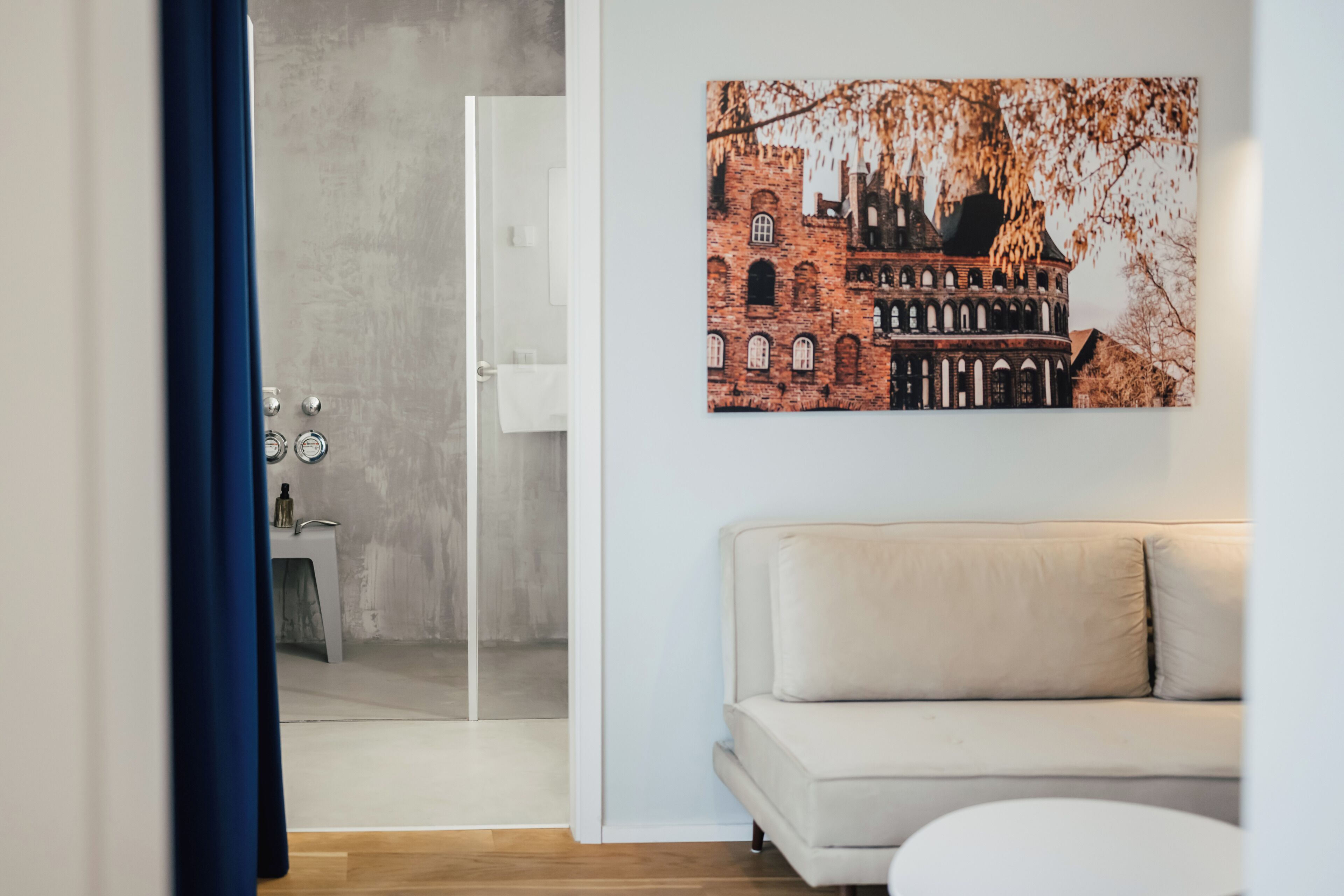 Uma sala de estar moderna com um sofá bege, uma porta de vidro que dá para um banheiro e um quadro com uma vista urbana na parede.