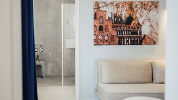 Uma sala de estar moderna com um sofá bege, uma porta de vidro que dá para um banheiro e um quadro com uma vista urbana na parede.