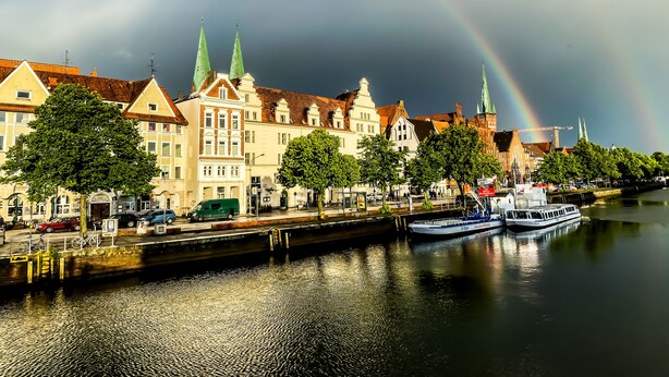 A riverside view with historic buildings, a boat docked on the water, and a rainbow in the sky.