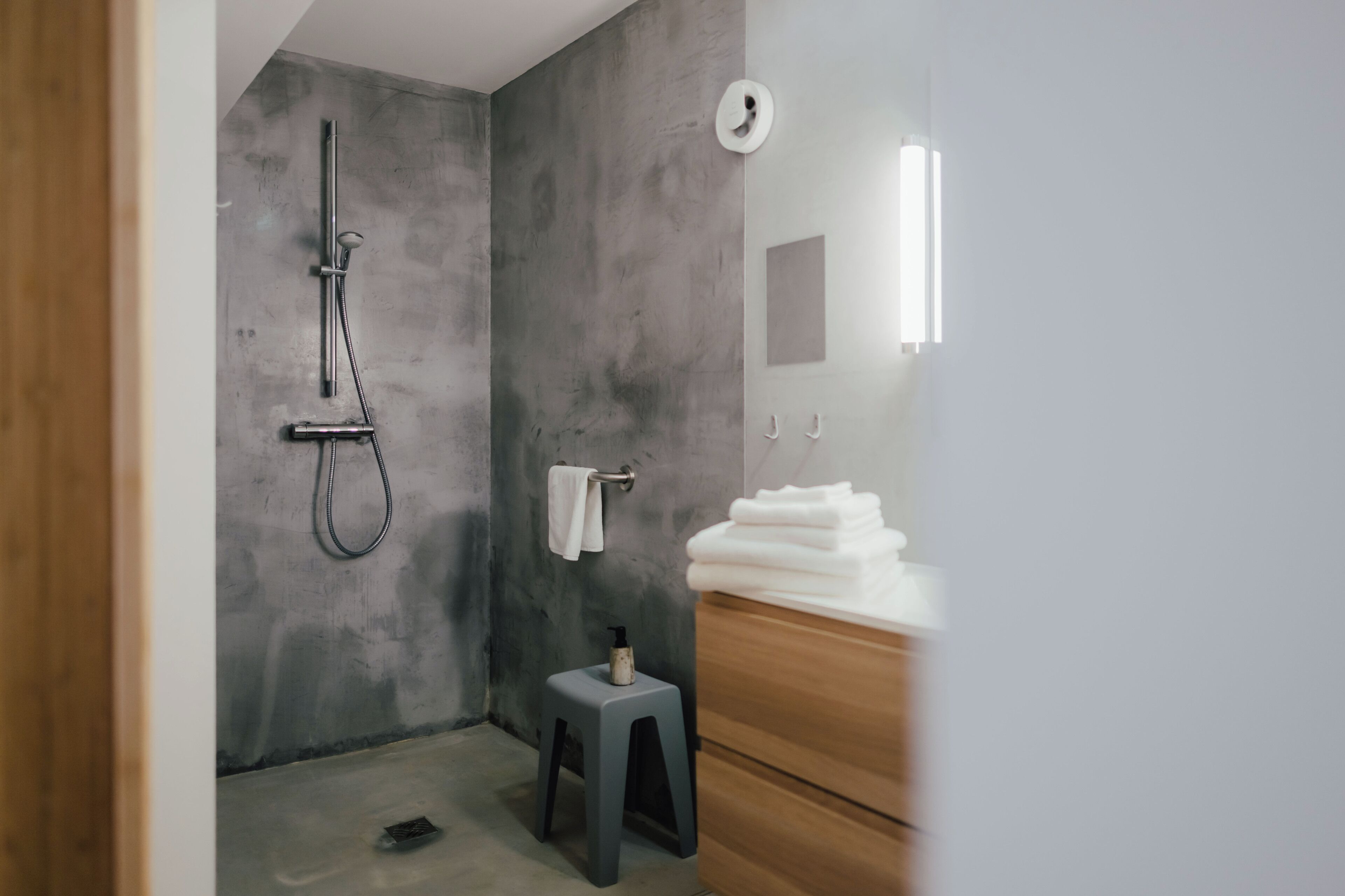 A modern bathroom with a concrete wall, a shower, a small stool, and a wooden vanity with towels.