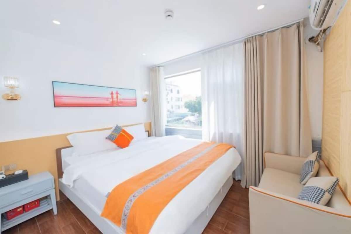 Una habitación de hotel con cama, mesita de noche, sofá, ventana con cortinas y un cuadro en la pared.