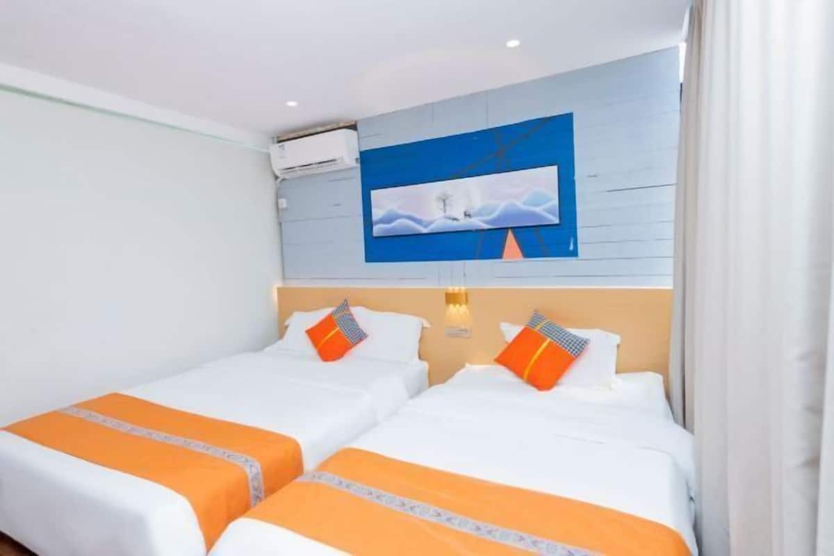 Habitación de hotel con dos camas, cada una con ropa de cama blanca y detalles naranja, un cuadro grande en la pared y un aire acondicionado instalado en el techo.