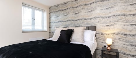 A bedroom with a bed, a nightstand, a lamp, and a window.