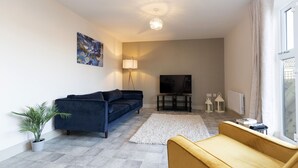 Ein Wohnzimmer mit einem dunkelblauen Sofa, einem gelben Sessel, einem weißen Teppich, einer Topfpflanze, einem Flachbildfernseher und einer Deckenleuchte.