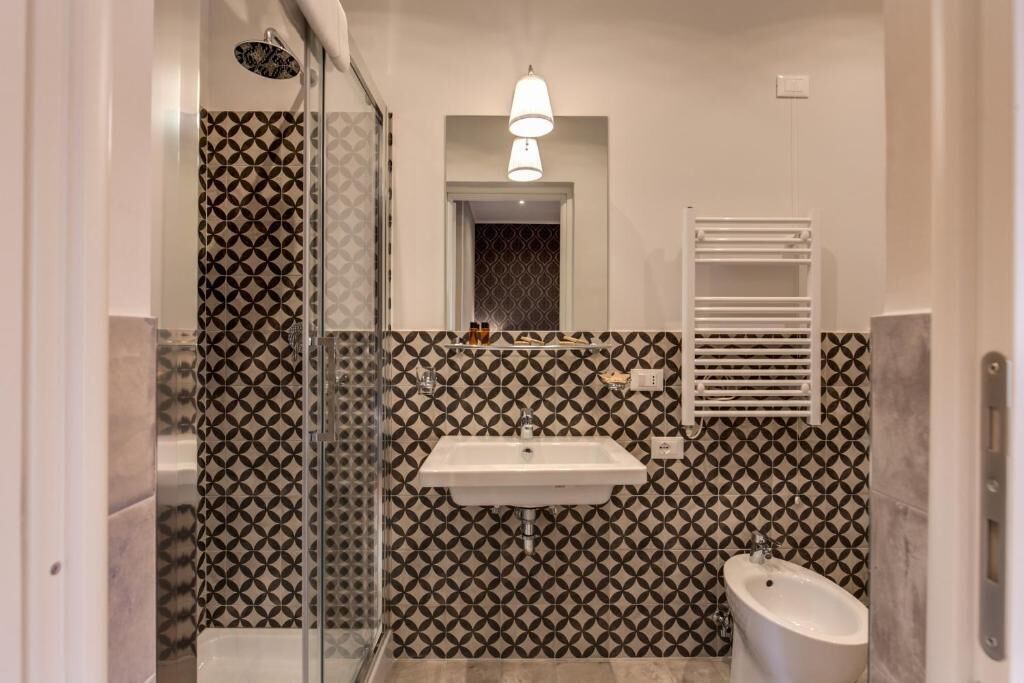 Un bany amb una paret de disseny geomètric, un lavabo blanc, un vàter i un box de dutxa de vidre.