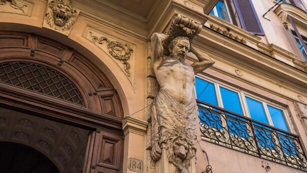 A building facade with a statue of a man holding a scroll, ornate architectural details, and a balcony with a wrought iron railing. -  magenta collection navona (Rome)