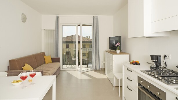 A modern kitchen with a stove, a dining table with two wine glasses and a bowl of chips, a sofa with a yellow pillow, and a sliding glass door leading to a balcony. - Appartamenti Alle Mura (Lazise)