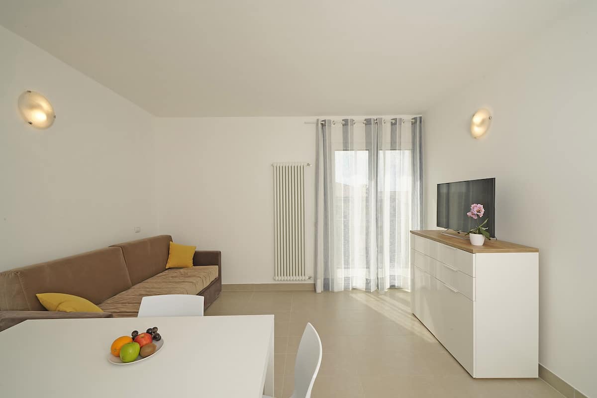 A modern living room with a sofa, a dining table with fruit, a TV on a cabinet, and a window with curtains.