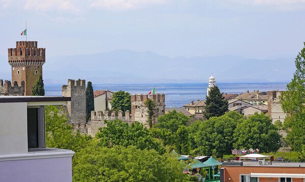 A historic town with medieval architecture, including a tower and a church with a bell tower, overlooking a body of water and mountains. - Appartamenti Alle Mura (Lazise)