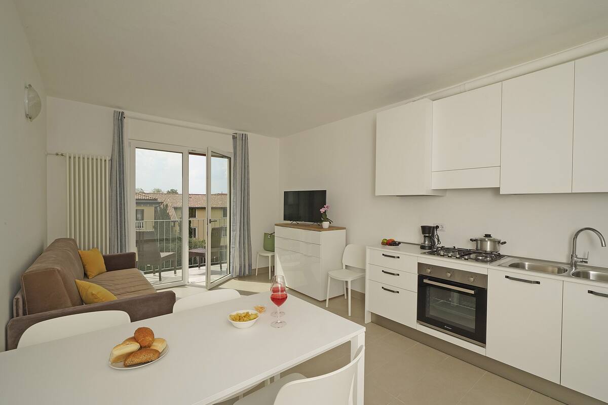 A modern kitchen with white cabinets, a dining table with a bowl of fruit and a glass of red wine, and a sofa with yellow cushions in a bright living area with large windows.