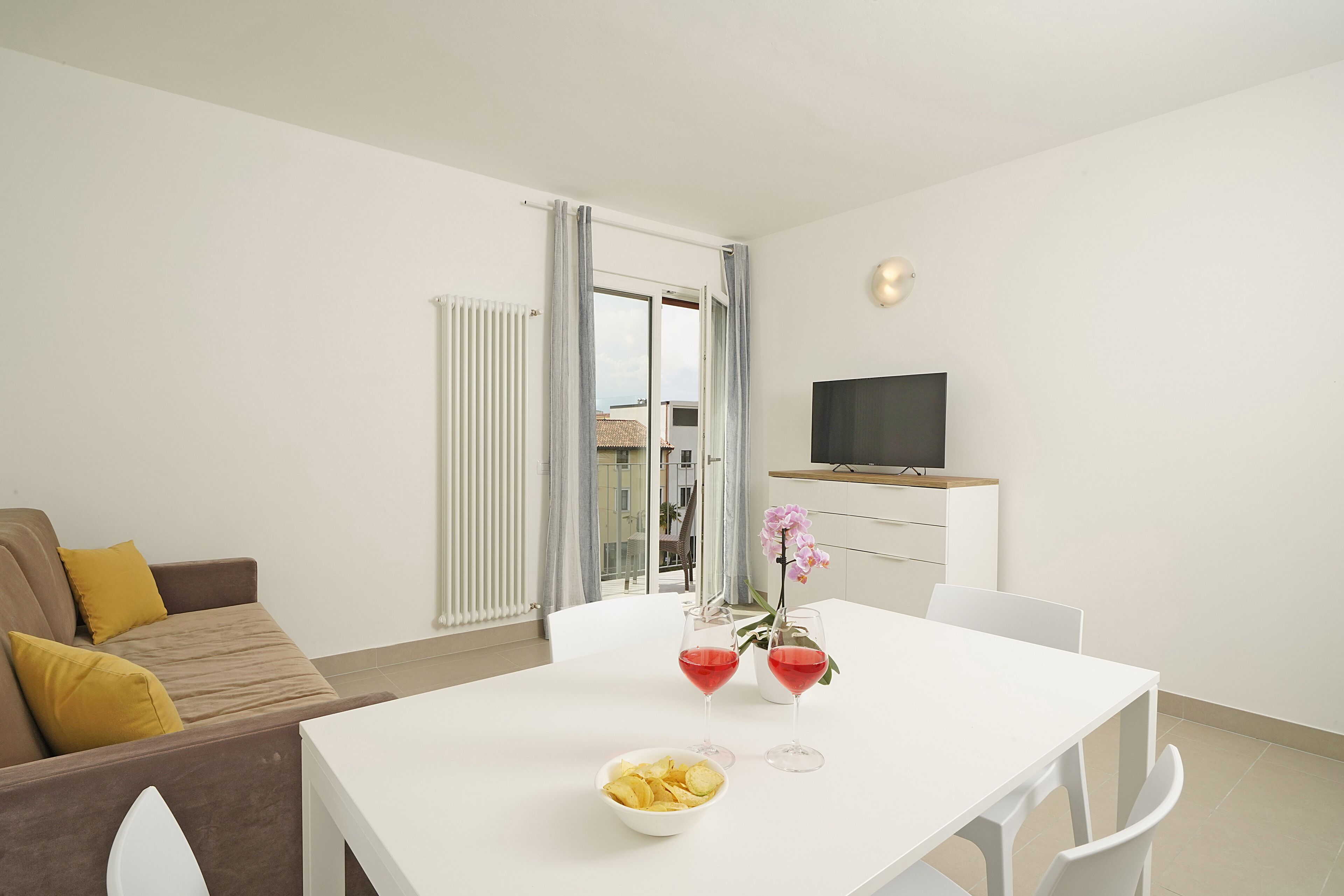 Una sala moderna con un sofá blanco, una mesa de comedor con dos bebidas rojas y un tazón de papas fritas, y un televisor montado en la pared.