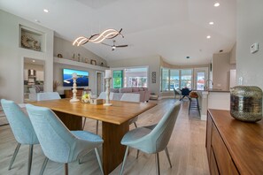 Un comedor amplio con una mesa de madera y sillas azul claro, un televisor en la pared y vistas al exterior a través de grandes ventanales.