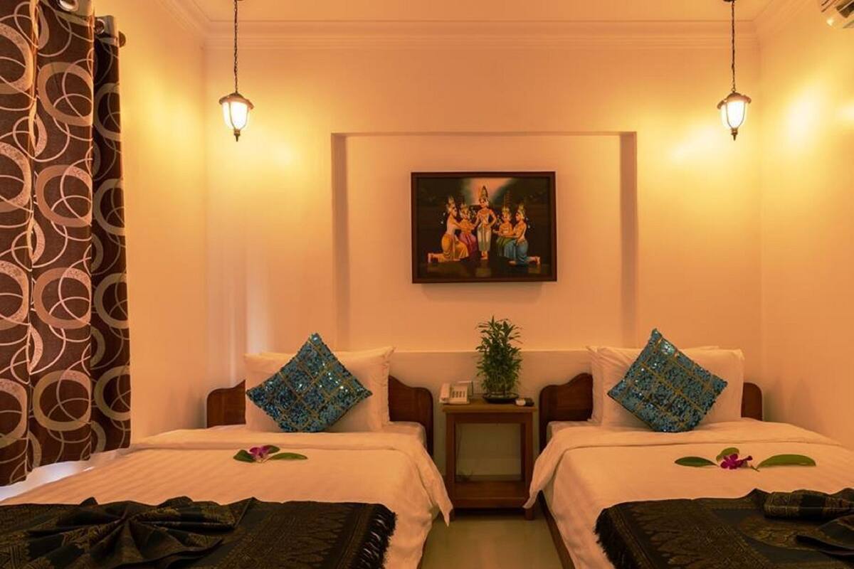 Une chambre d’hôtel avec deux lits, un tableau au mur, une table de chevet avec une plante et un rideau à motifs.