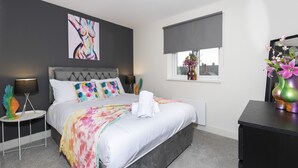 Een moderne slaapkamer met een grijs bekleden hoofdbord, een witte spreidek en een kleurrijk plaid. Er is een raam met uitzicht op gebouwen, een nachtkastje met een lamp en een decoratieve vaas met bloemen.