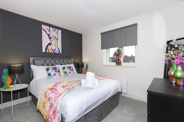 A modern bedroom with a grey upholstered headboard, a white bedspread, and a colorful throw blanket. There is a window with a view of buildings, a bedside table with a lamp, and a decorative vase with flowers. - 4 Avery Gardens - 1 Bedroom Apartment (Darlington)