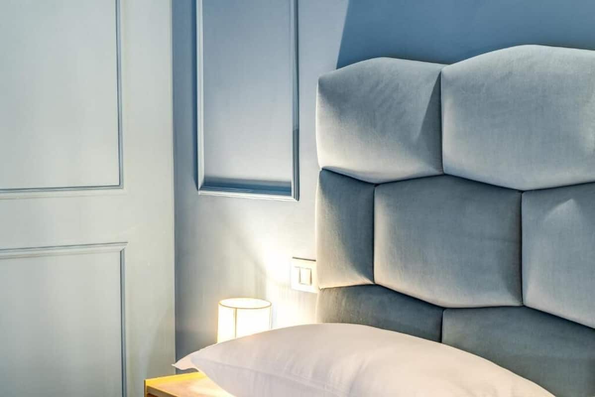 A modern bedroom with a tufted headboard, a bedside table with a lamp, and a window providing natural light.