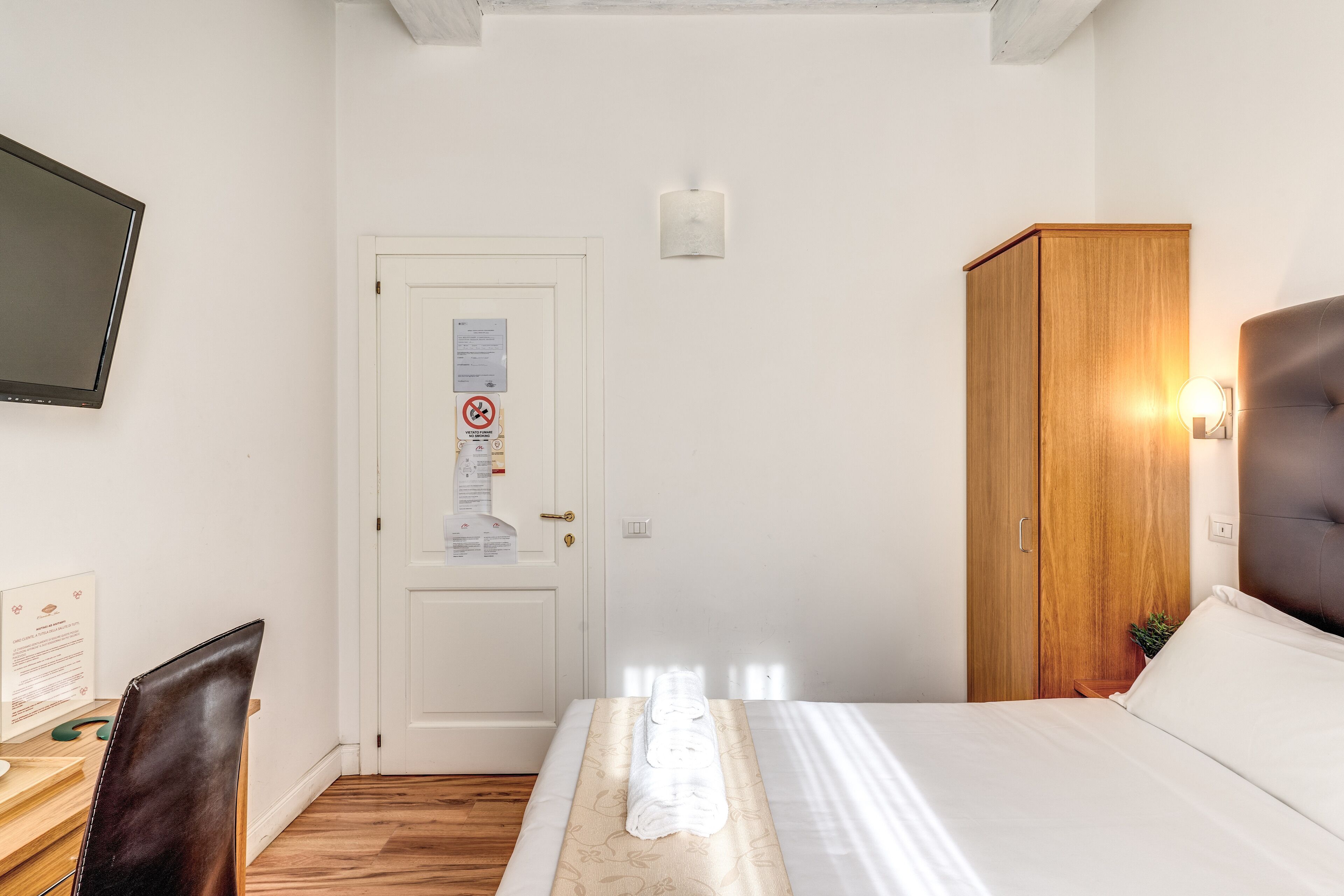 Habitación de hotel con cama, escritorio y silla, armario, televisor y puerta con un aviso.