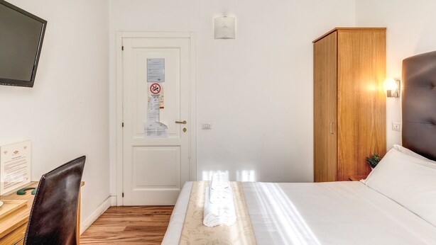 A hotel room with a bed, a desk with a chair, a wardrobe, a television, and a door with a notice. - Magenta Collection Cancello (Rome)