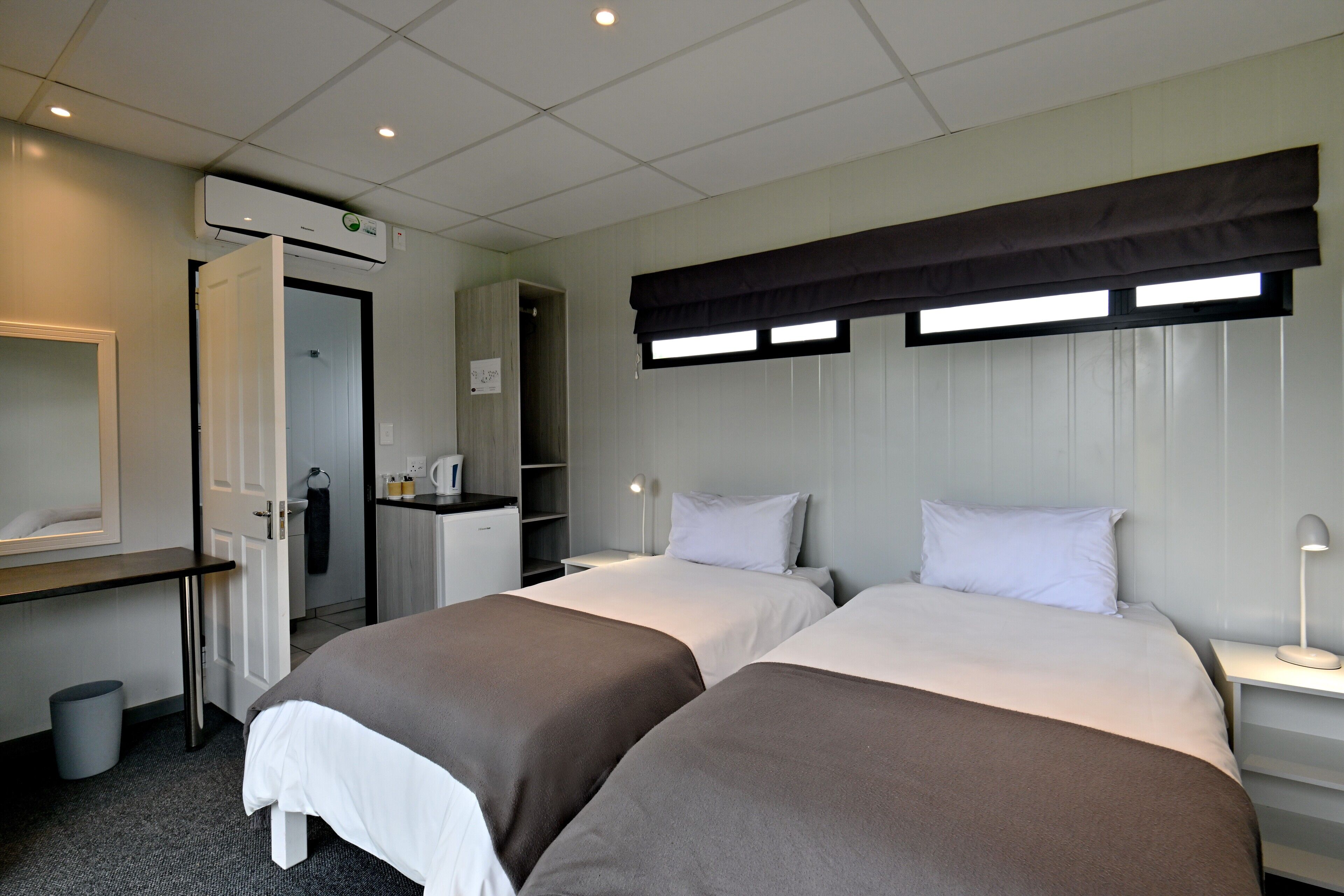 Une chambre d’hôtel avec deux lits, un bureau, une chaise, une poubelle et une fenêtre avec des stores.