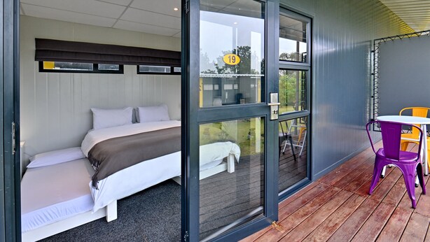 A room with a bed, a window with a number '19', and a patio area with a table and chairs. - SleepOver Phabeni Gate (Mbombela)