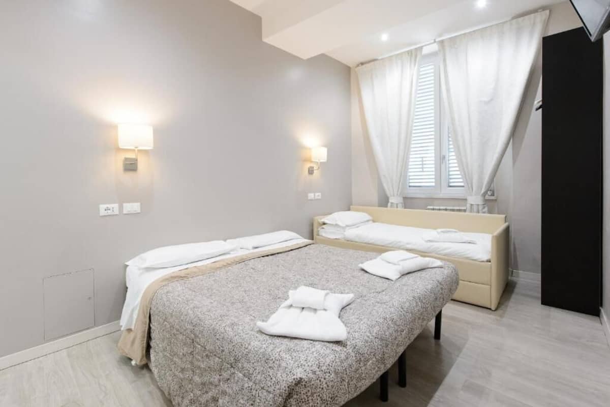 A modern hotel room with two beds, a large window with curtains, and wall-mounted lighting.