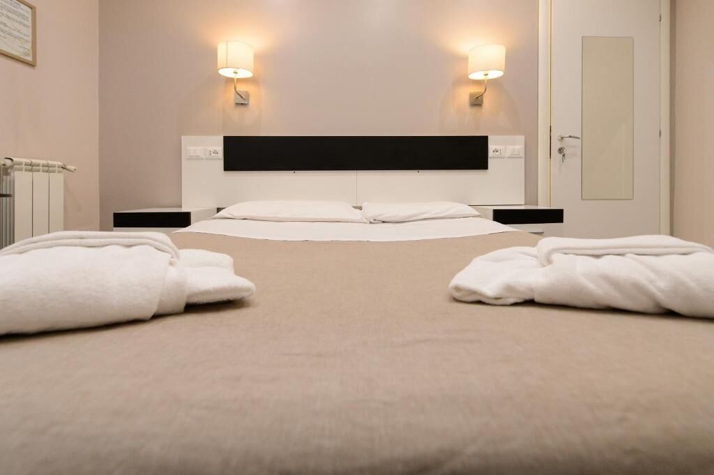 A hotel room with a bed, two bedside tables, a nightstand, a wall-mounted lamp, and a mirror.