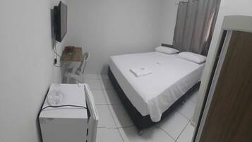 Quarto de hotel pequeno e limpo, com uma cama, escrivaninha, cadeira e televisão na parede.