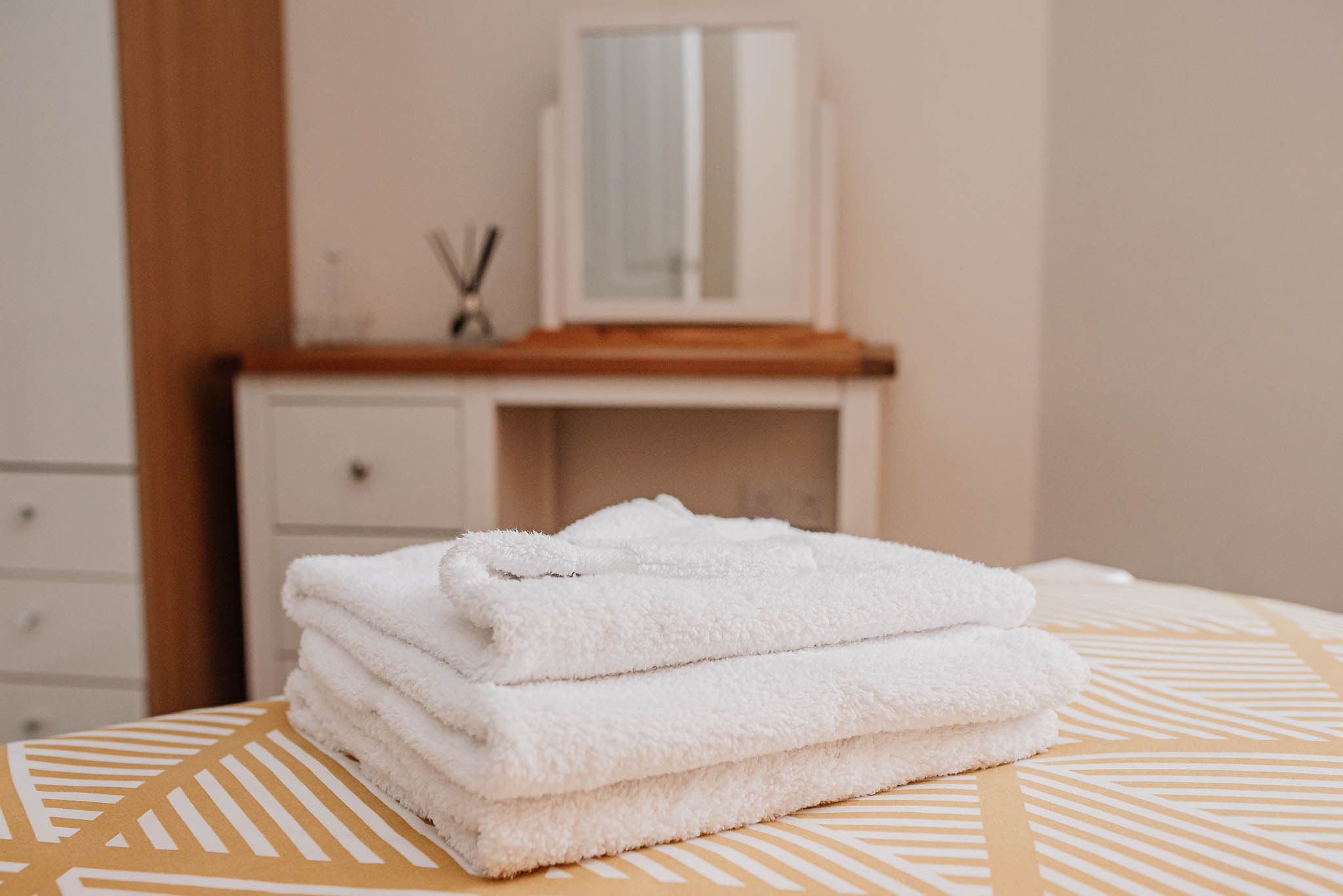 A neatly folded stack of white towels on a bed with a geometric patterned bedspread.