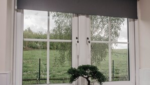 Une fenêtre donnant sur un champ verdoyant et des arbres, un pot noir contenant un bonsaï, et un cadre de fenêtre blanc.