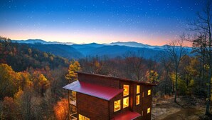 Eine zweistöckige Holzhütte mit vielen Fenstern, eingebettet in eine bewaldete Berglandschaft bei Nacht.