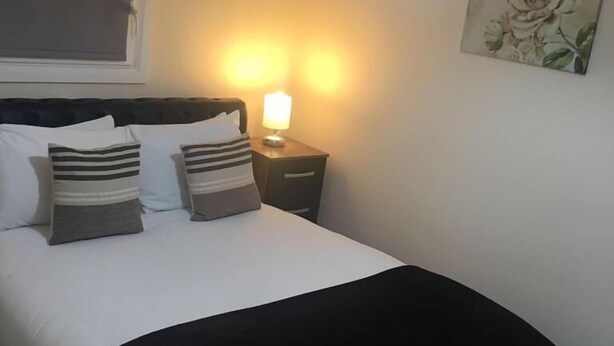 A neatly made bed with striped pillows, a bedside lamp, and a floral painting on the wall. - North Crescent (Leeds)