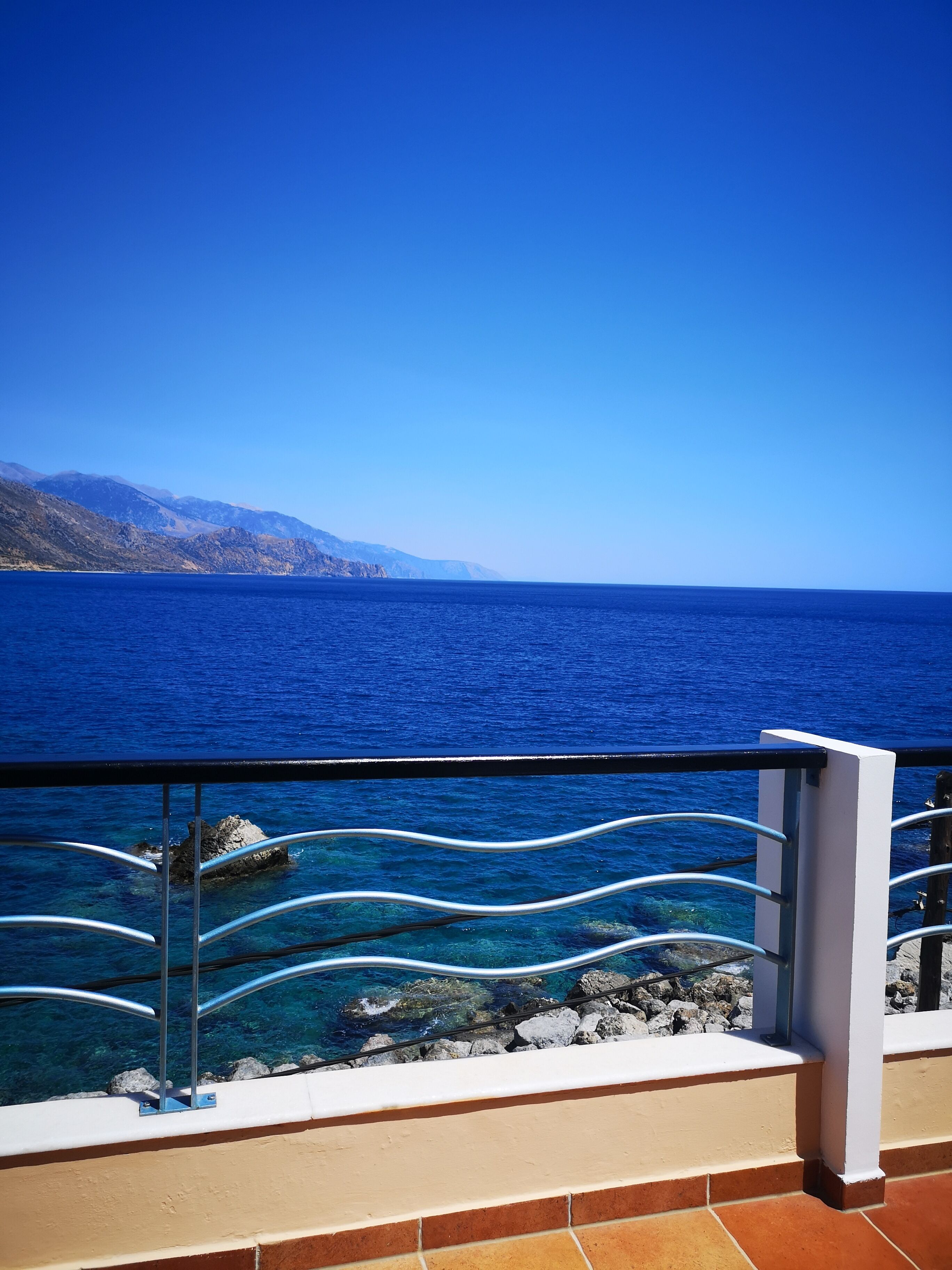 A balcony with a metal railing overlooking a clear blue sea and distant mountains.