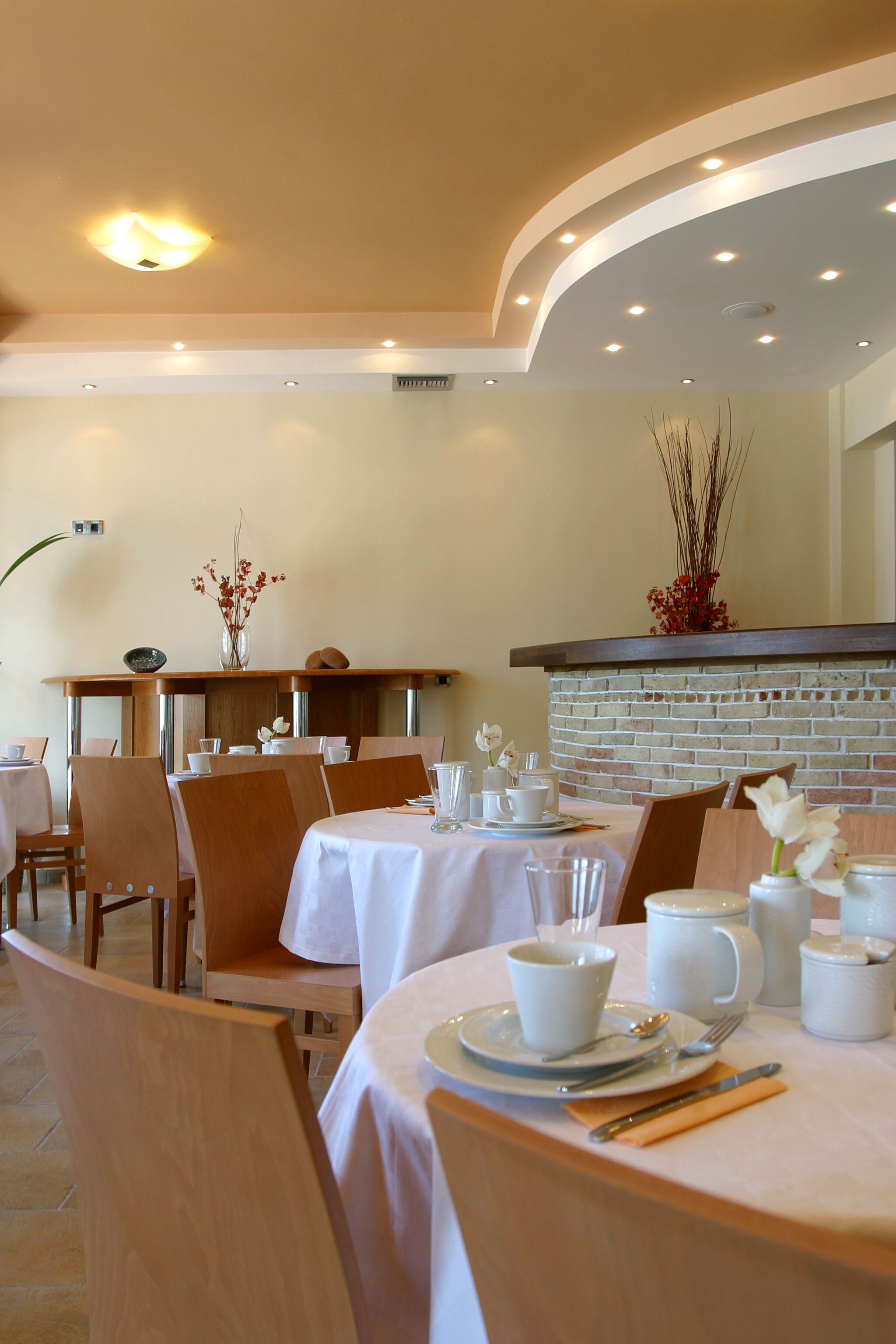 Un espace repas avec des tables rondes, des chaises et des nappes blanches. On y trouve des vases remplis de fleurs et, au fond, un mur en briques.