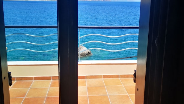 A view from a room with a tiled floor and a door leading to a balcony overlooking a body of water and distant mountains. - Hotel On The Rocks (Kantanos-Selino)