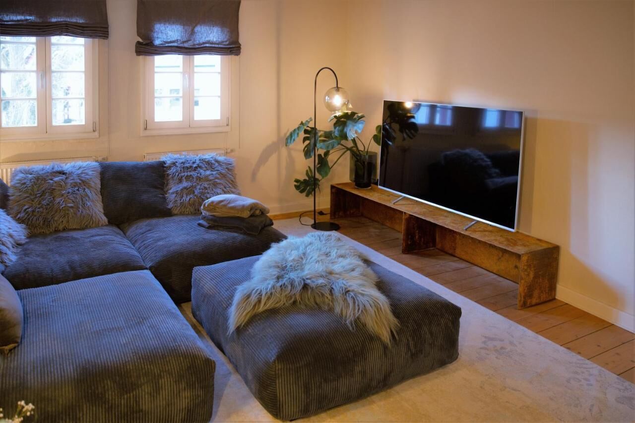 Een woonkamer met een hoekbank, een flatscreen televisie, een houten bankje en een plant in pot.